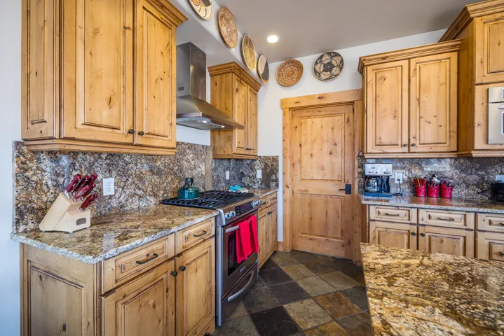 Kitchen featuring gas range, wall chimney range hood, dark stone counters, stone tile flooring, and brown cabinetry