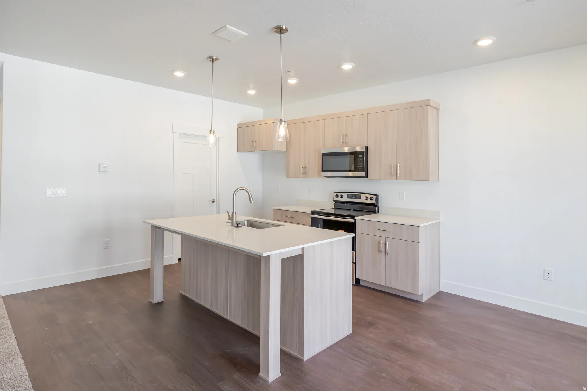 Kitchen featuring light wood finish cabinets, stainless steel appliances, a kitchen island with sink, pendant lighting, and dark wood-type flooring