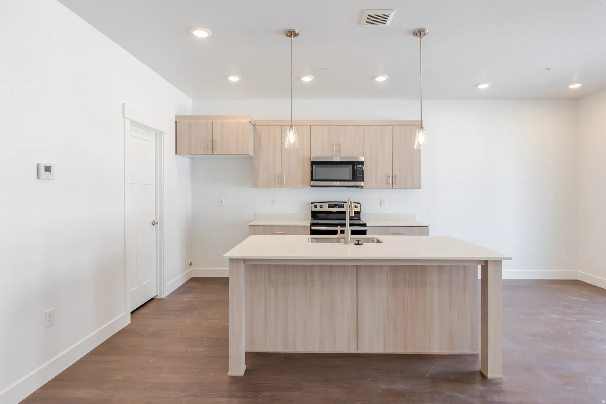 Kitchen featuring light wood finish cabinetry, an island with sink, hanging light fixtures, stainless steel appliances, and dark wood-style floors