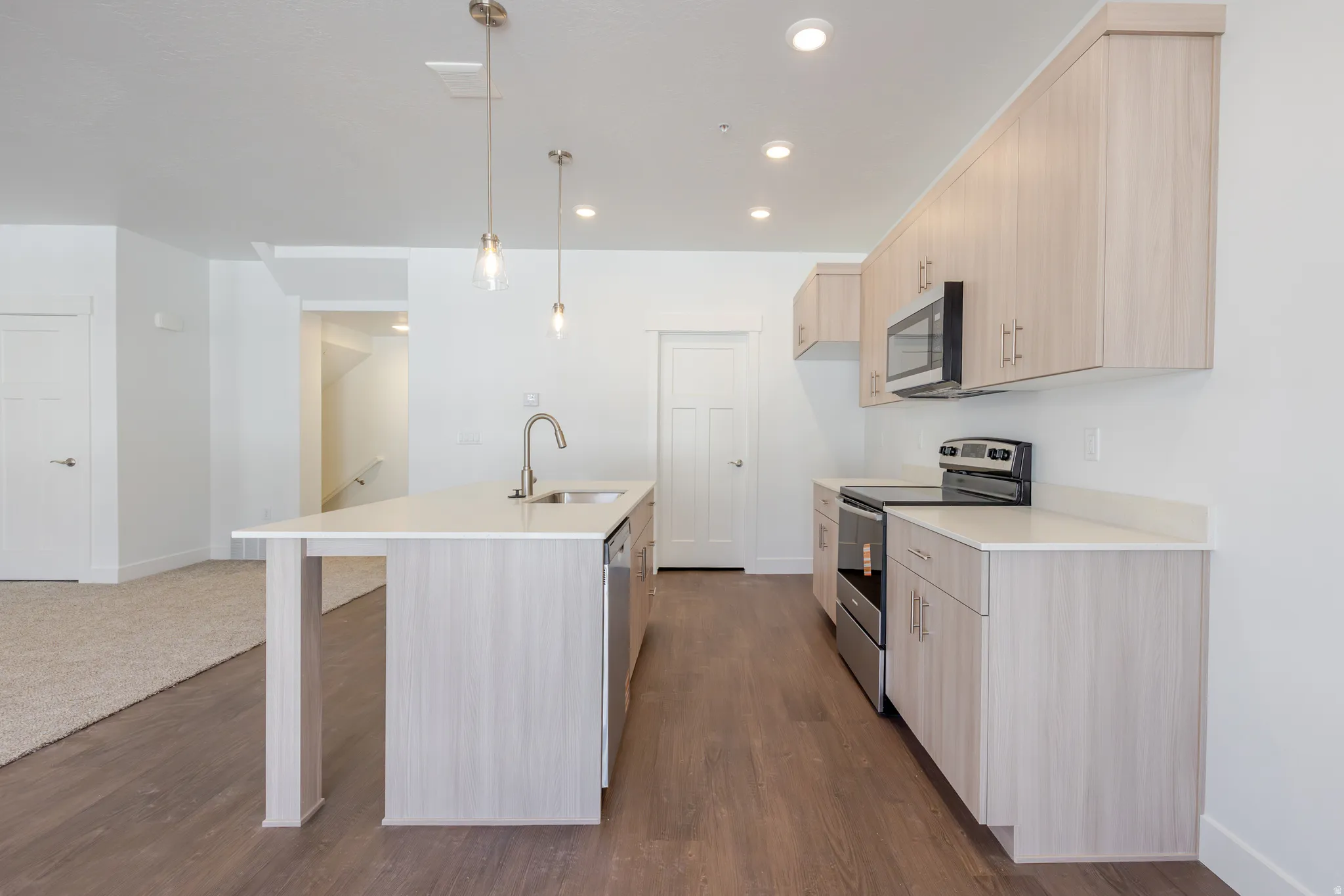 Kitchen with stainless steel appliances, light wood finish cabinetry, an island with sink, dark wood-type flooring, and pendant lighting