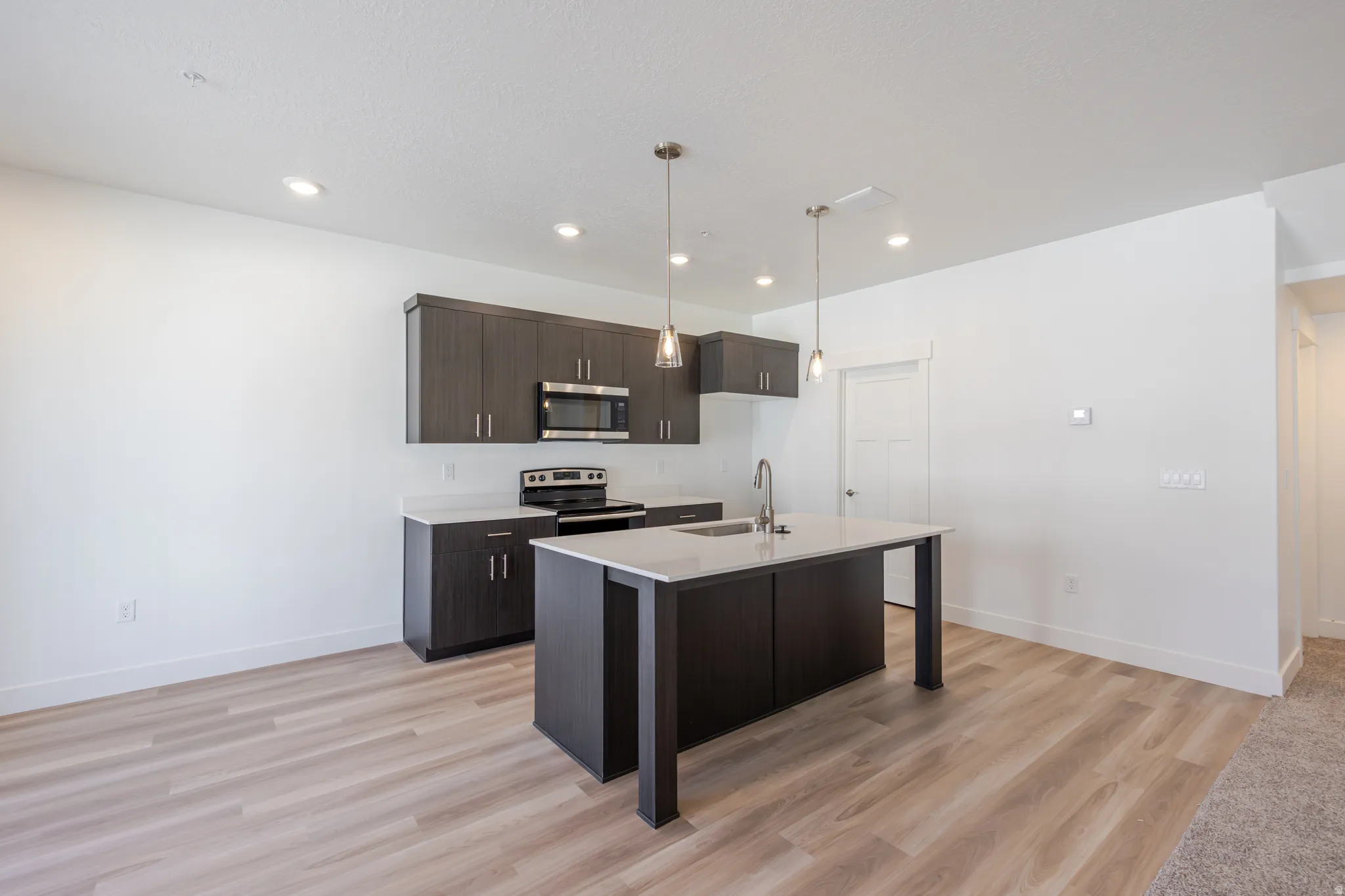 Kitchen with dark wood finish cabinetry, stainless steel appliances, hanging light fixtures, light wood-style flooring, and an island with sink