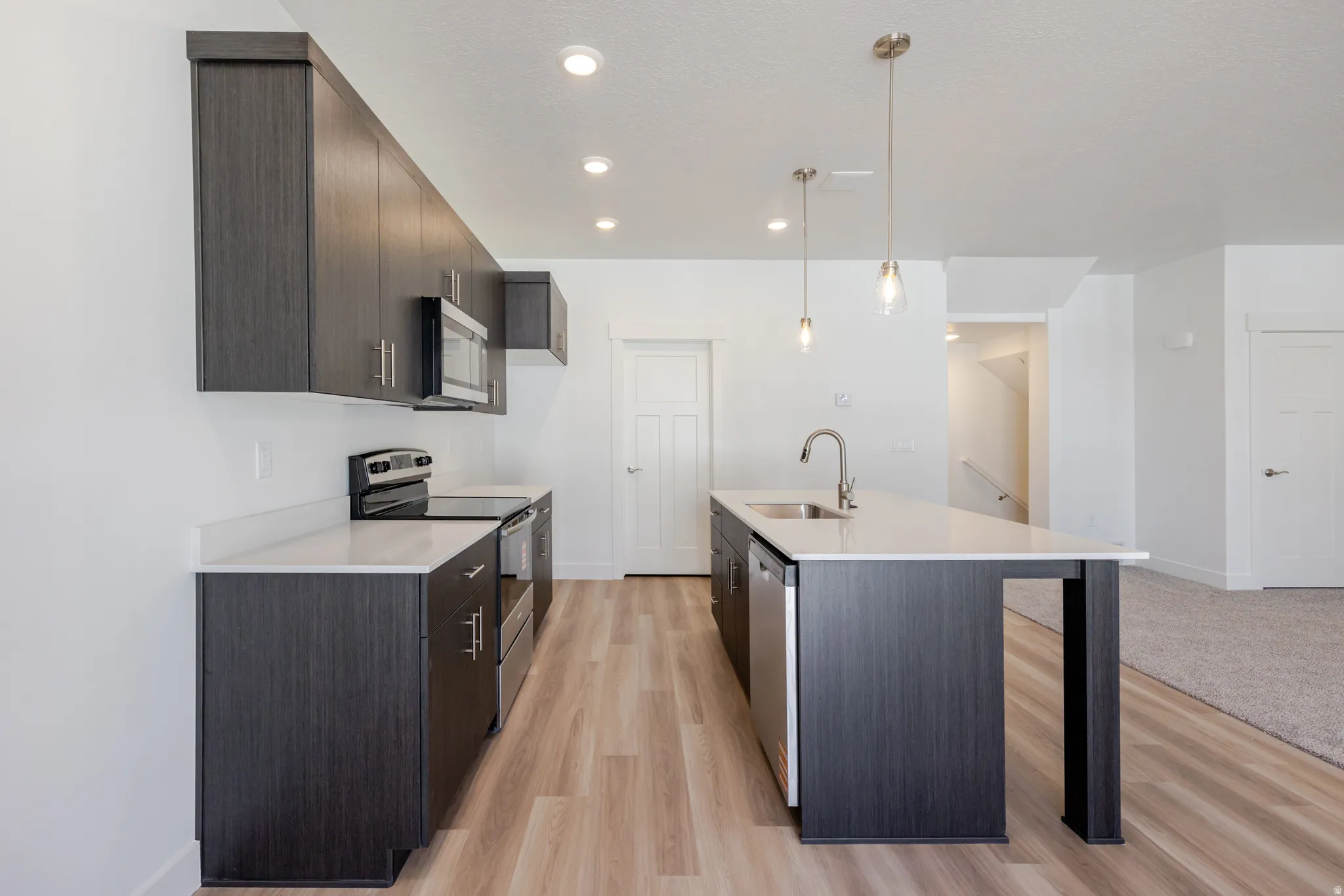 Kitchen featuring stainless steel appliances, a center island with sink, decorative light fixtures, dark wood finish cabinets, and light wood-style flooring
