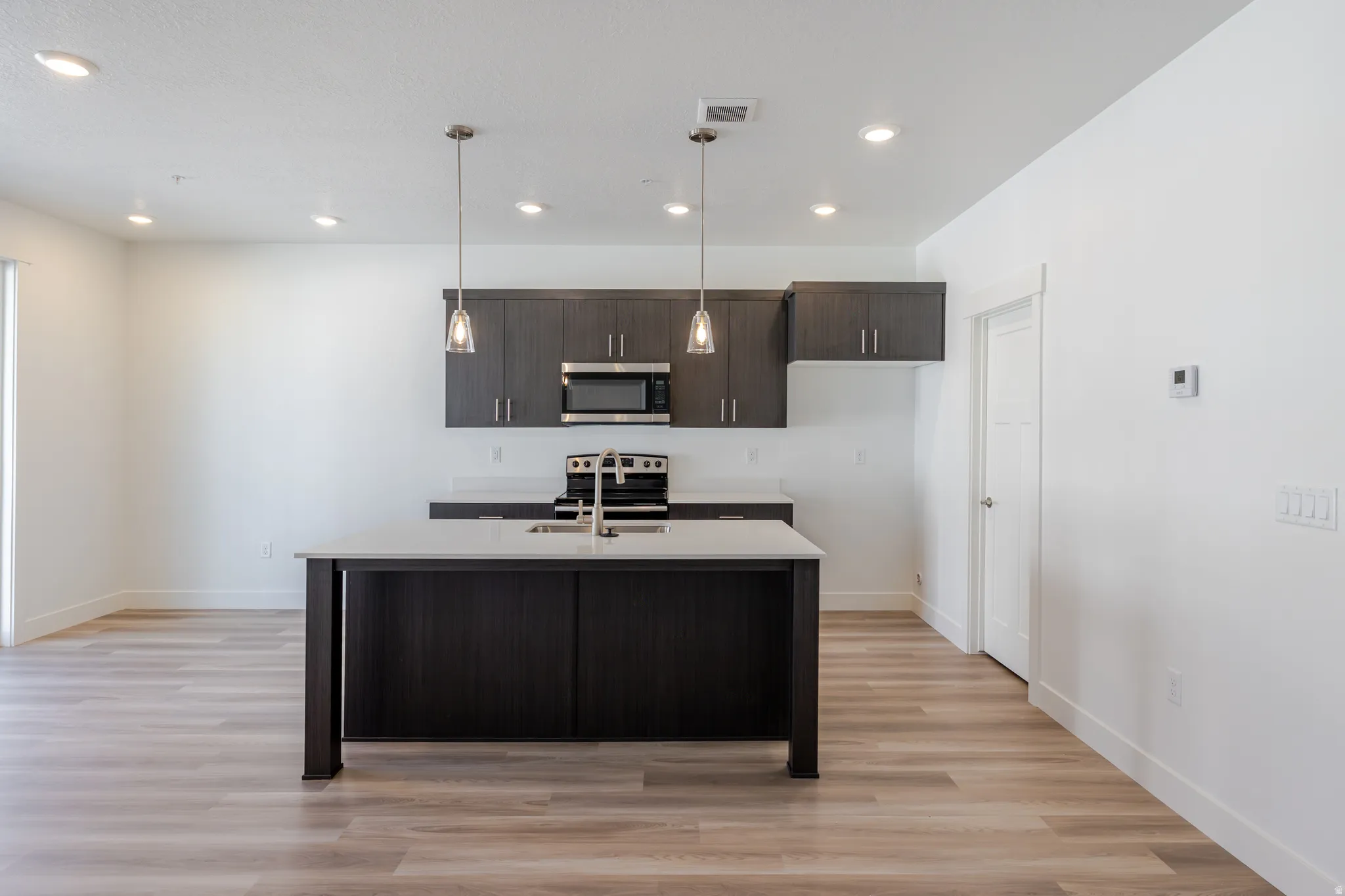 Kitchen featuring dark wood finish cabinetry, light wood-style floors, stainless steel appliances, an island with sink, and pendant lighting