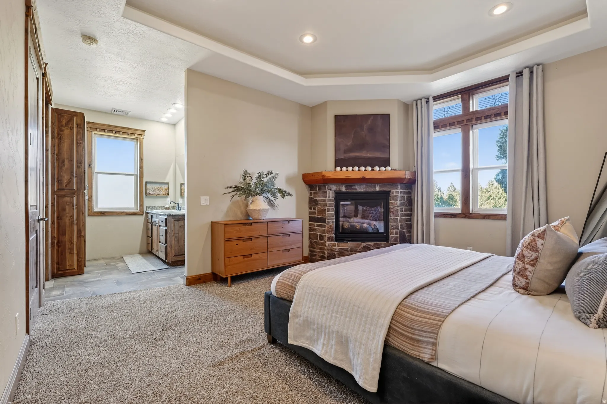 Bedroom with a tray ceiling, a glass covered fireplace, light colored carpet, and connected bathroom