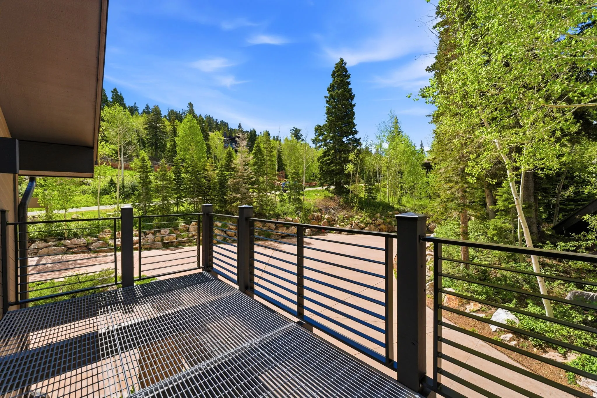 Outdoor metal grating deck featuring a black horizontal bar railing, surrounded by mature deciduous and evergreen trees under a clear sky