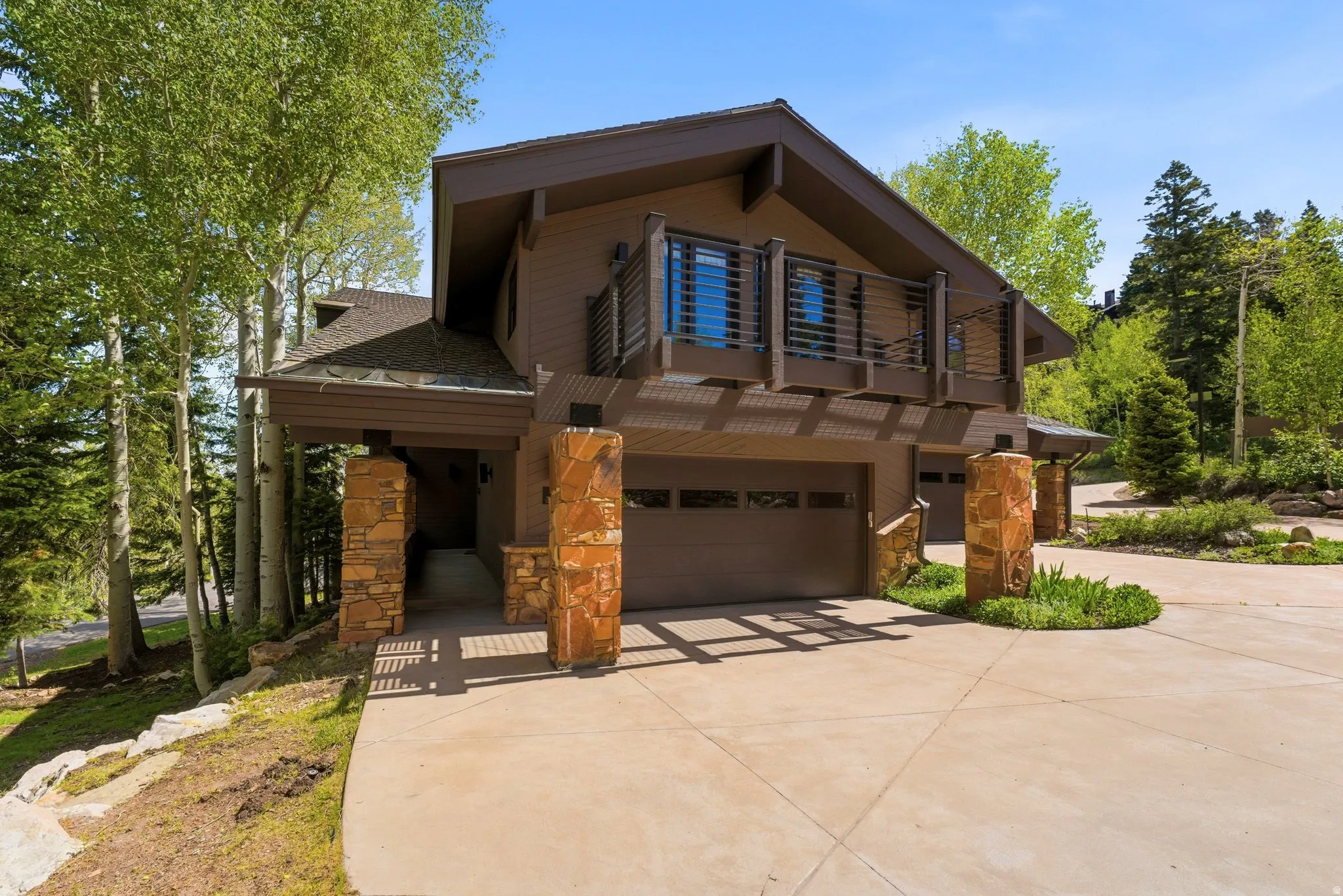 Contemporary brown exterior with dual garage doors, stone columns, upper-level balcony with horizontal railings, and a concrete driveway