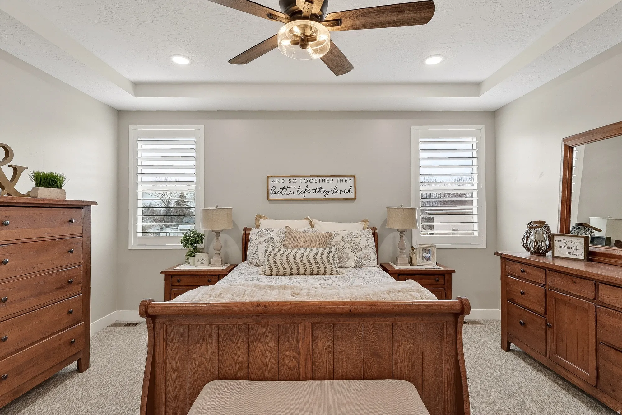 Bedroom with light colored carpet, multiple windows, ceiling fan, and a tray ceiling