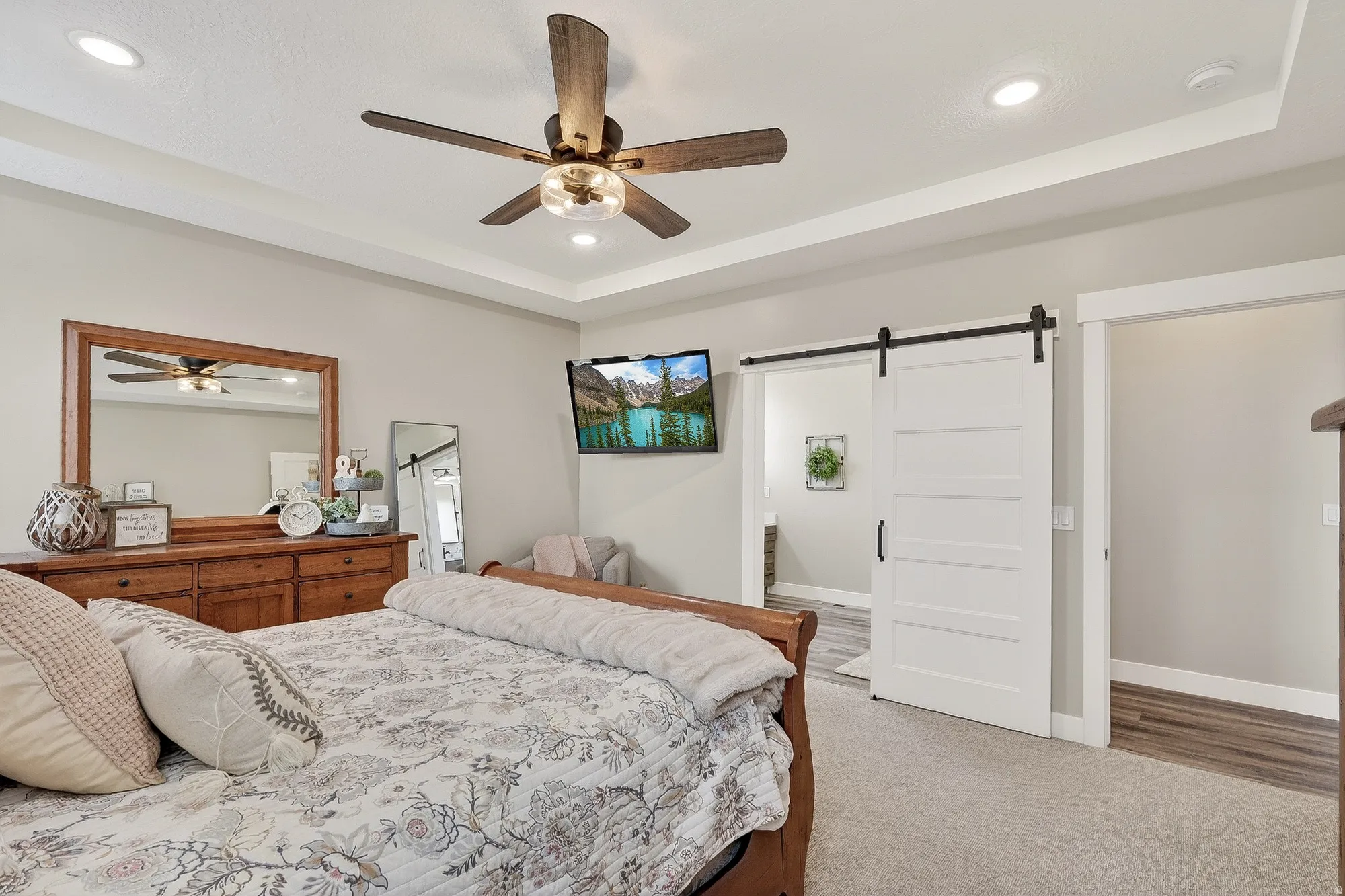 Carpeted bedroom with a barn door, a tray ceiling, ensuite bath, recessed lighting, and a ceiling fan