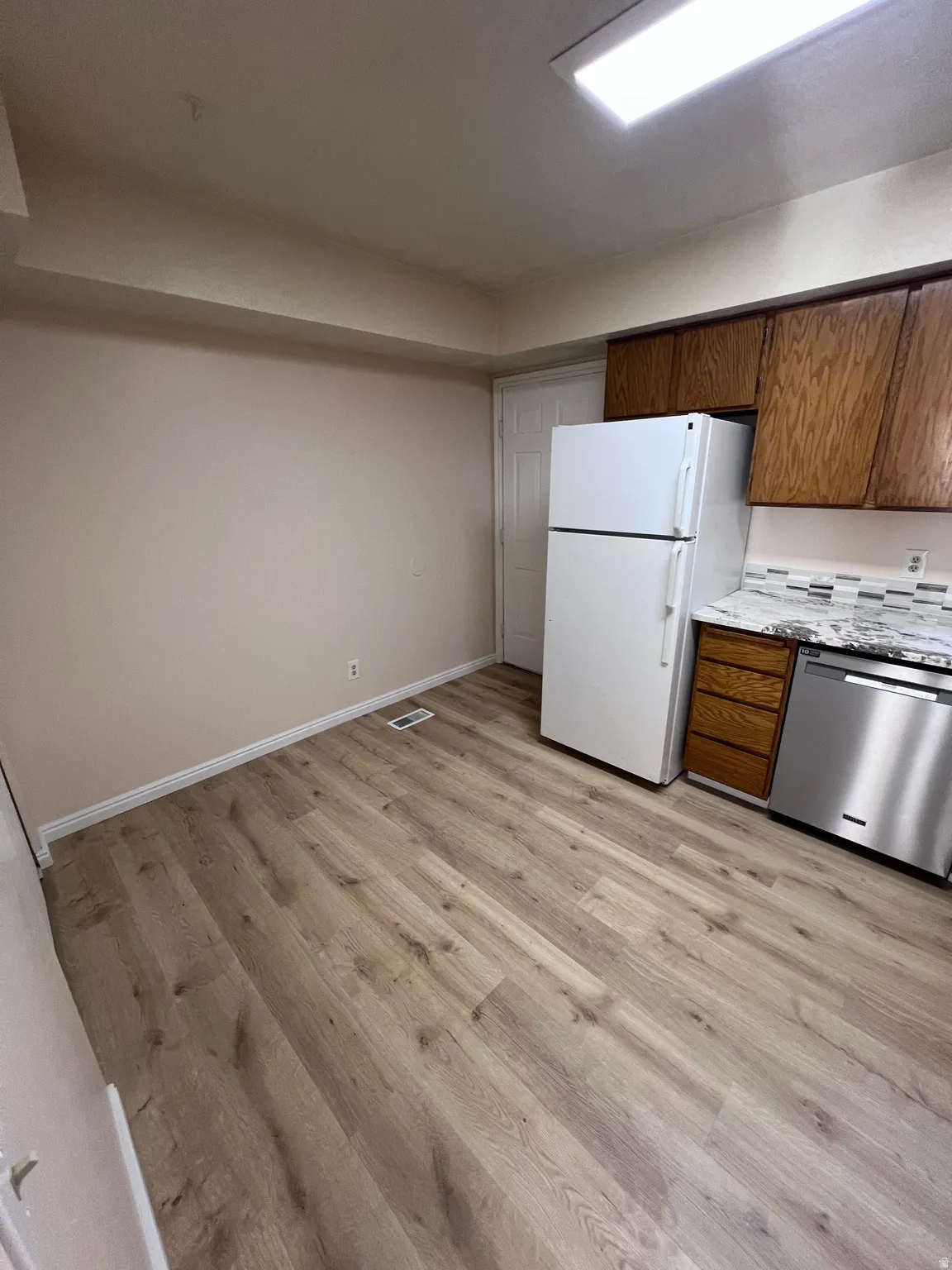 Kitchen with stainless steel dishwasher, brown cabinets, freestanding refrigerator, light wood-style flooring, and light stone countertops