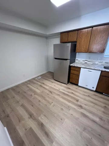 Kitchen featuring dishwasher, freestanding refrigerator, light wood-type flooring, light stone counters, and brown cabinetry