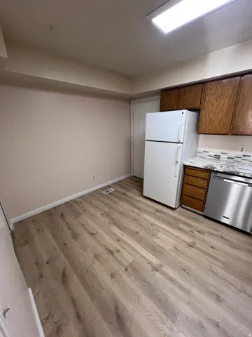 Kitchen with stainless steel dishwasher, brown cabinets, freestanding refrigerator, light wood-style flooring, and light stone countertops