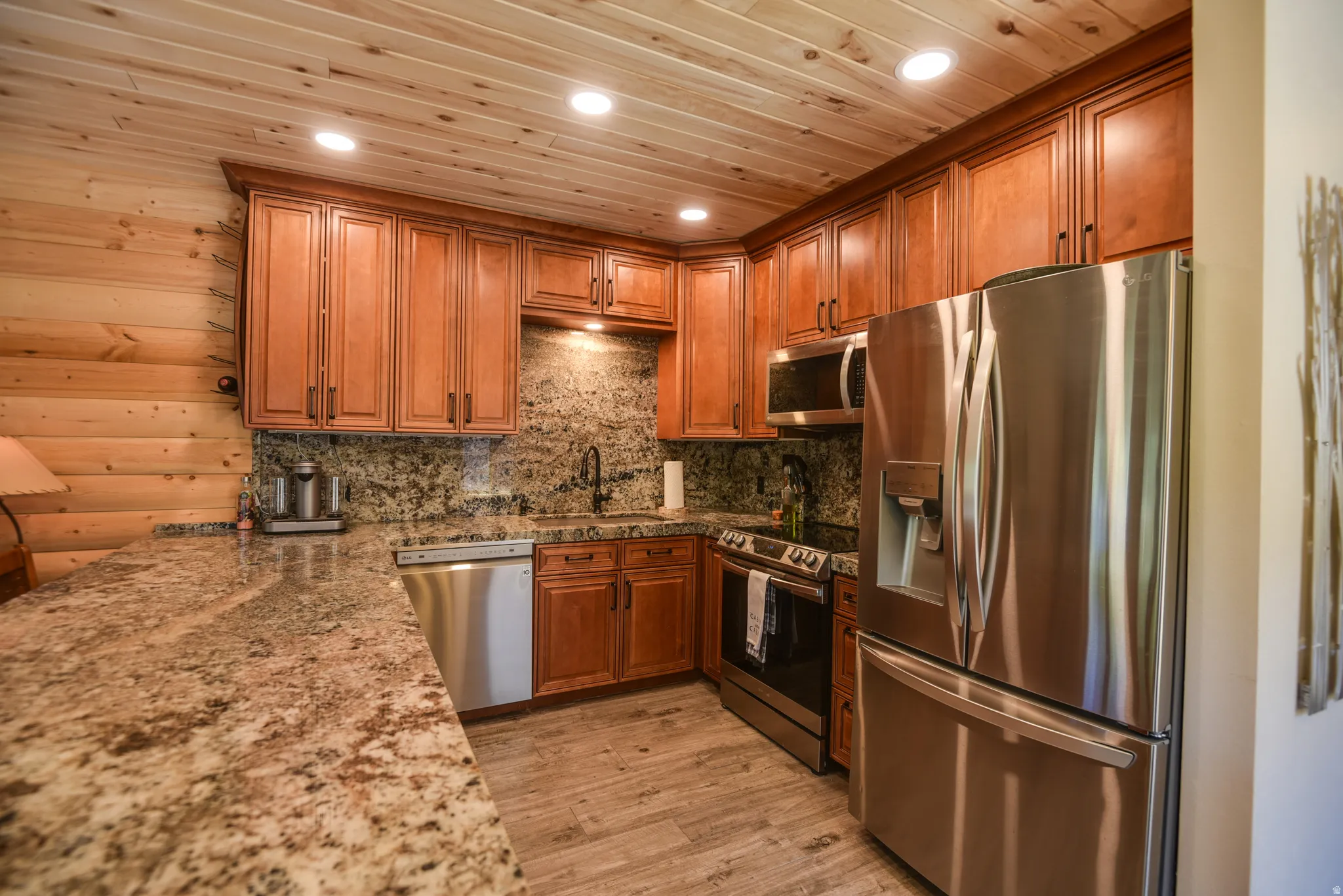 Kitchen with appliances with stainless steel finishes, brown cabinetry, light stone countertops, light wood-style flooring, and tasteful backsplash
