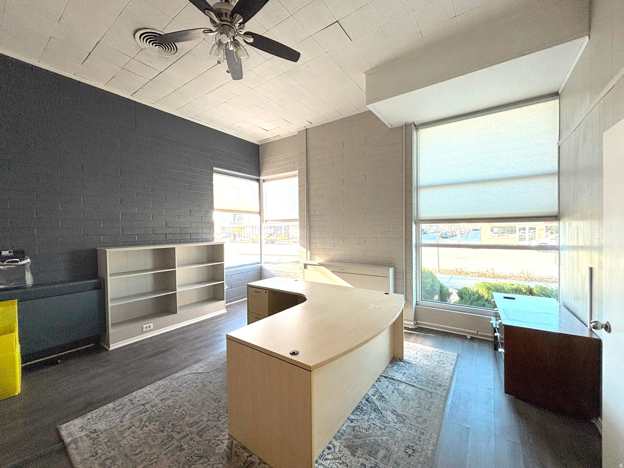 Office area featuring dark wood-style flooring, ceiling fan, and brick wall