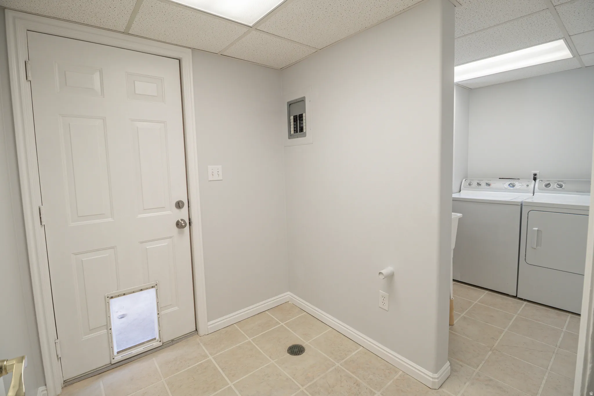 Washroom with light tile patterned floors, a paneled ceiling, washer and dryer, and electric panel