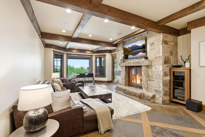 Stone fireplace with an integrated television niche, wood-finish ceiling beams, recessed lighting, expansive windows, and geometric pattern tile flooring