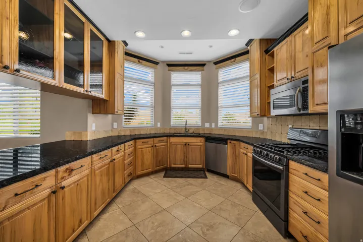 Kitchen with appliances with stainless steel finishes, dark stone counters, brown cabinetry, glass insert cabinets, and recessed lighting