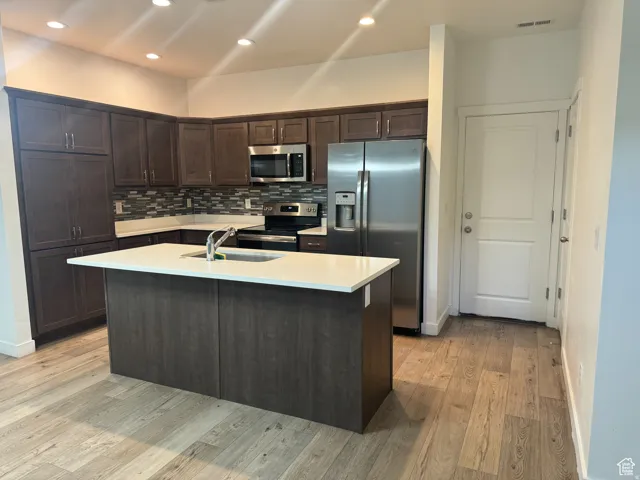 Kitchen with dark brown cabinetry, stainless steel appliances, tasteful backsplash, a center island with sink, and recessed lighting