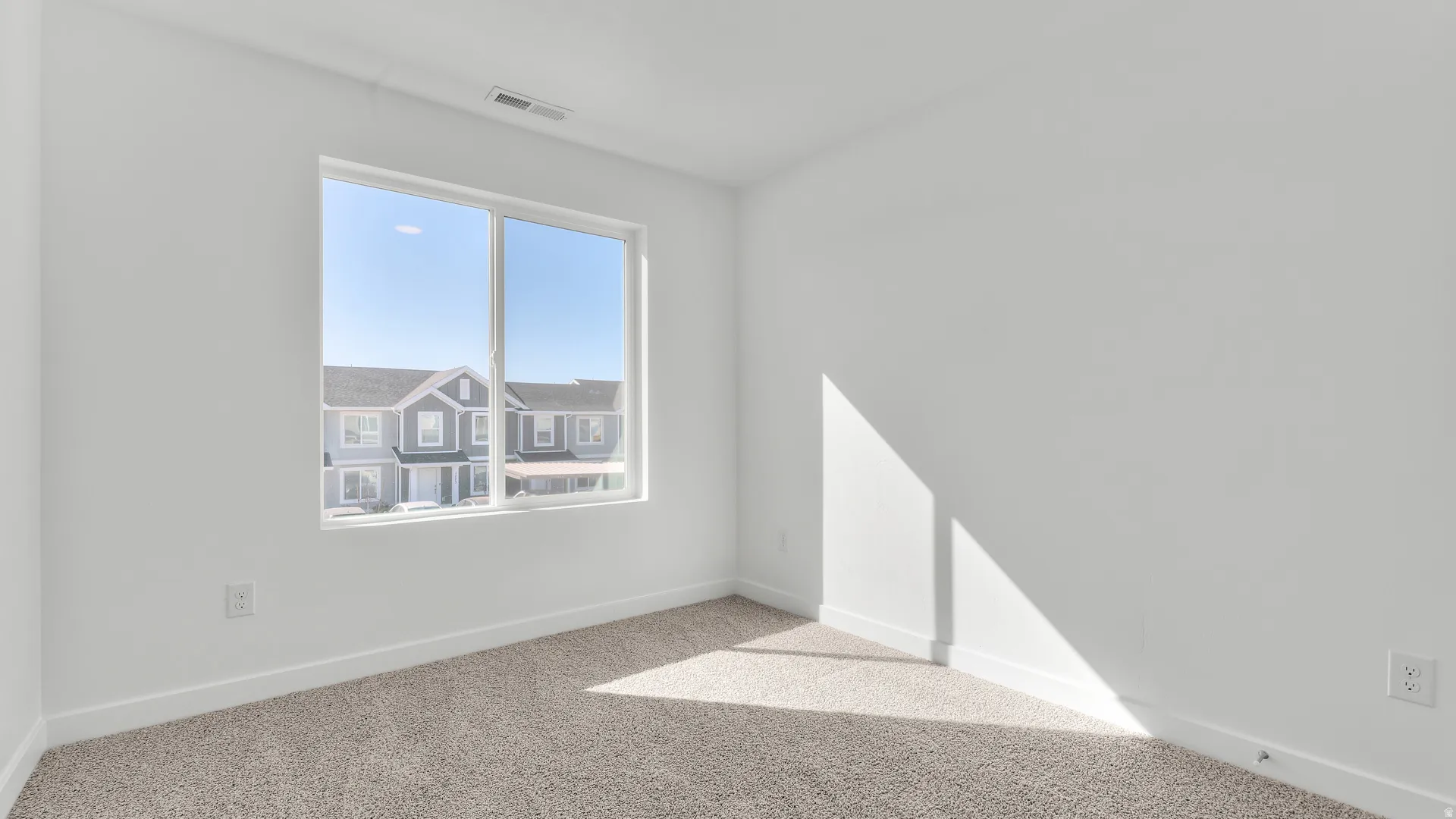 Bright interior space featuring a large window, soft neutral carpeting, white baseboards, and a white ceiling with a visible vent