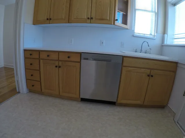 Apt # 8 > Kitchen featuring stainless steel dishwasher, light countertops, glass insert cabinets, light flooring, and brown cabinets
