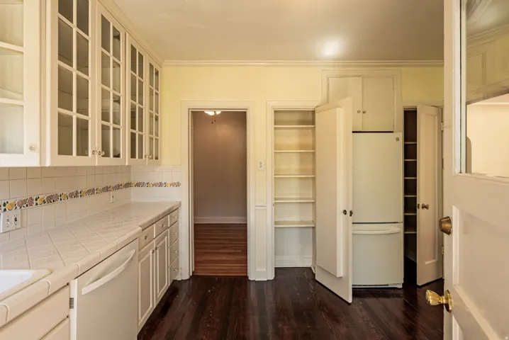 Kitchen featuring white cabinetry, glass-front upper cabinets, white tile countertops, white tile backsplash with decorative border, and dark wood-finish flooring