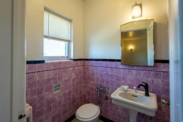 Bathroom featuring purple wall tiling with black accent trim, a pedestal sink with a matte black faucet, a toilet, a window with blinds, and a wall-mounted mirror with an overhead light fixture