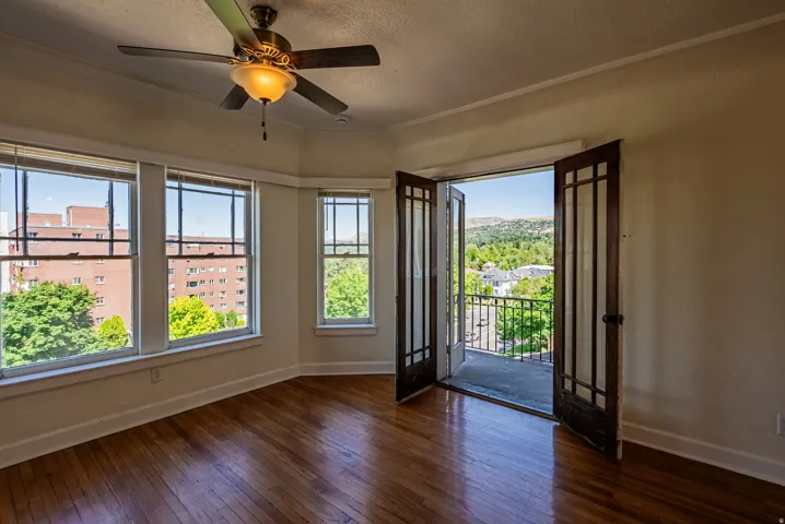 Hardwood flooring throughout featuring a corner bay window arrangement, a ceiling fan with integrated lighting, and double doors opening to a balcony with metal railings