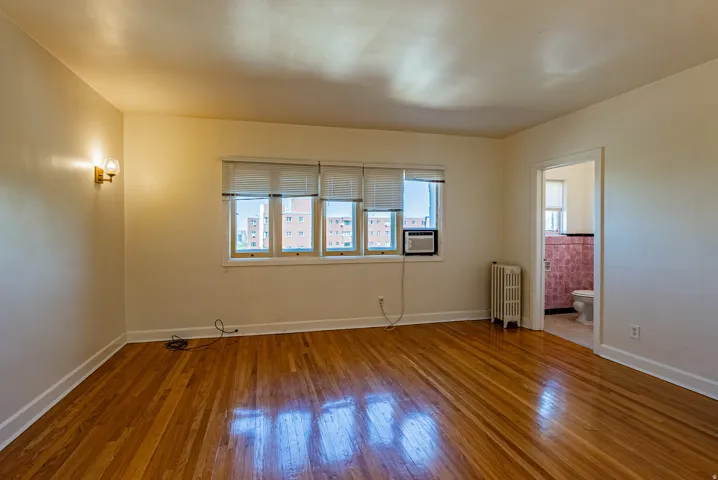 Spacious room featuring polished wood flooring, multiple windows with blinds, a wall-mounted light fixture, and a white baseboard heater