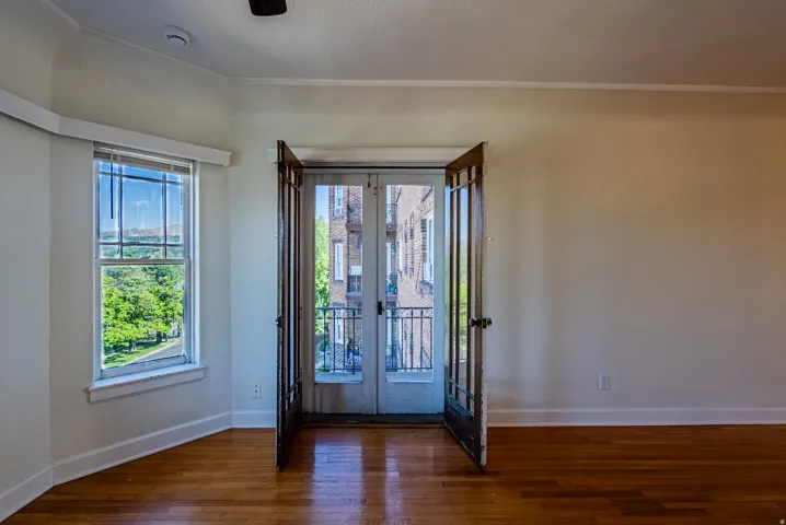 Wood-finish flooring throughout, featuring a bay window with natural light, a ceiling fan, and French doors opening to an exterior balcony with wrought iron railing
