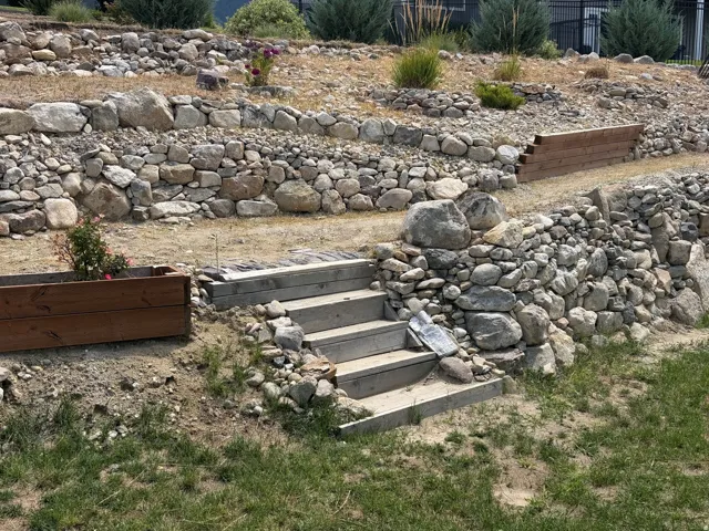 Tiered retaining walls constructed from natural stone and boulders, featuring integrated wood planking and concrete steps