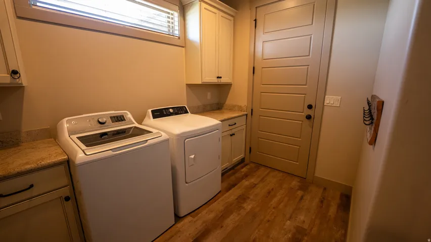 Dedicated laundry area featuring wood-finish flooring, light-toned cabinetry, solid surface countertops, and a horizontal window with blinds