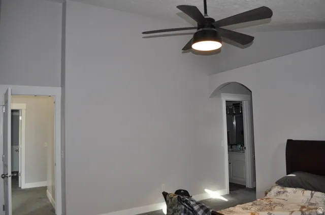 Bedroom with dark colored carpet, vaulted ceiling, arched walkways, ceiling fan, and ensuite bath