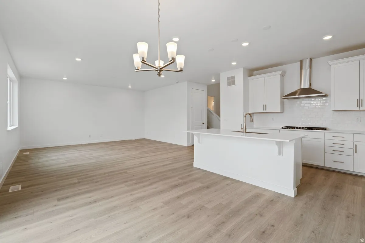 Kitchen with white cabinets, hanging light fixtures, a chandelier, a center island with sink, and wall chimney exhaust hood