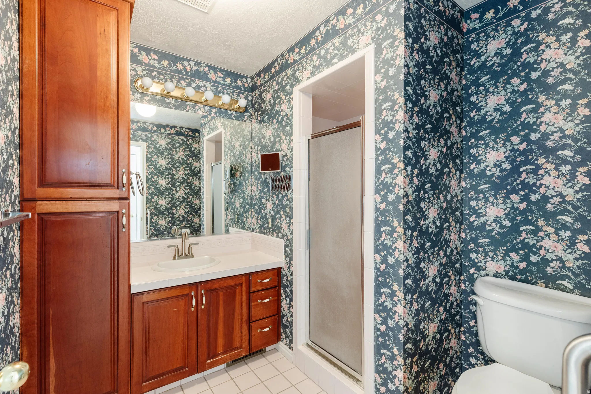 Bathroom with a shower stall, vanity, light tile patterned floors, wallpapered walls, and a textured ceiling