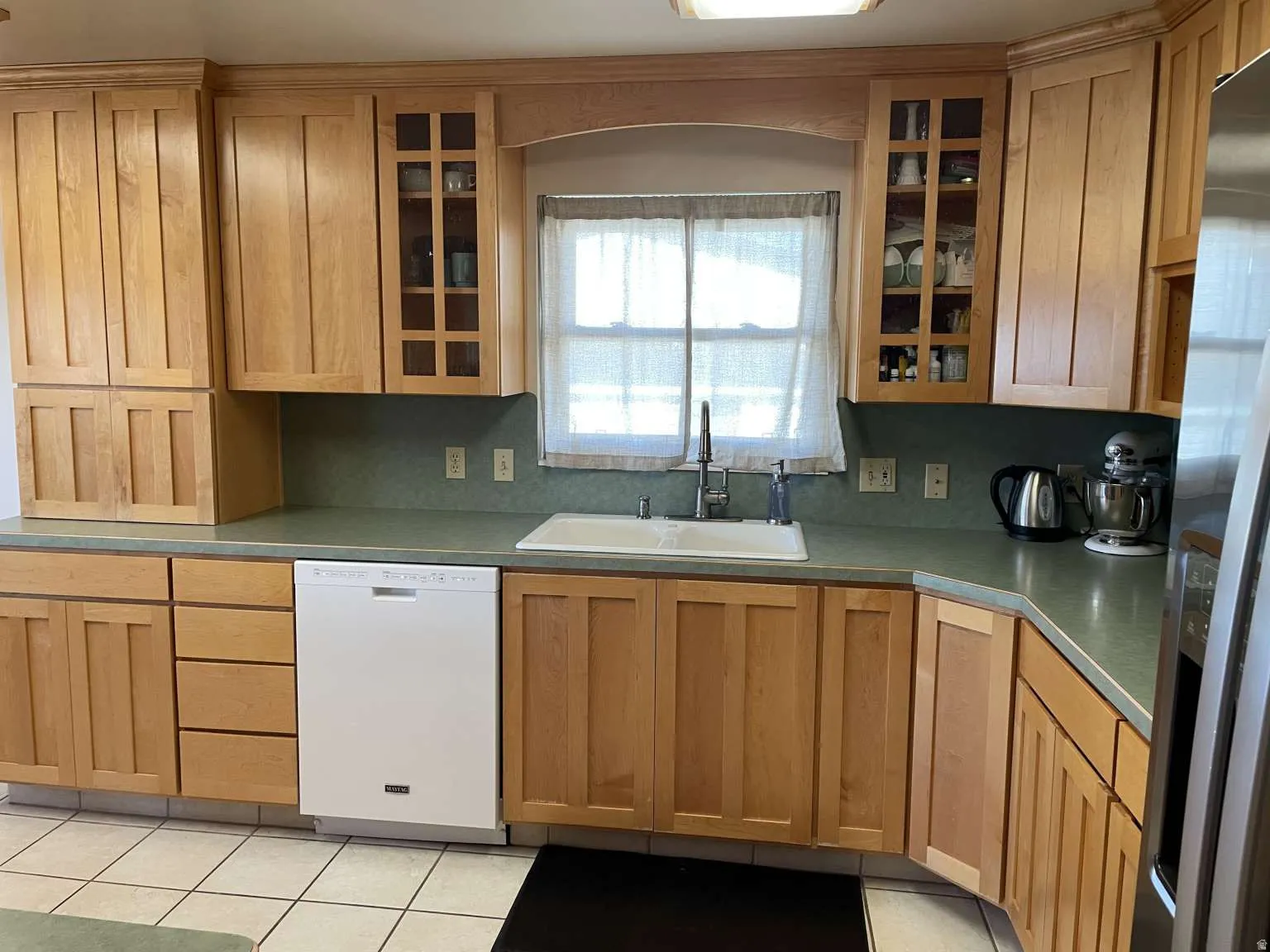 Kitchen with dishwasher, glass insert cabinets, stainless steel fridge, brown cabinets, and light tile patterned floors