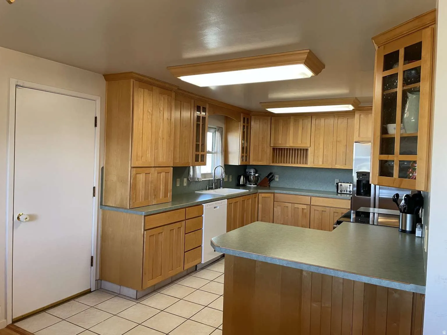 Kitchen featuring glass insert cabinets, a peninsula, white dishwasher, light tile patterned flooring, and brown cabinetry