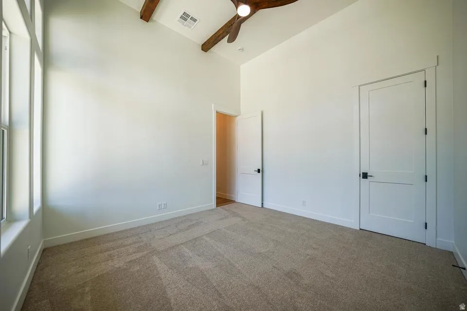 Spacious carpeted room featuring a vaulted ceiling with exposed wood beams, a ceiling fan, and multiple windows