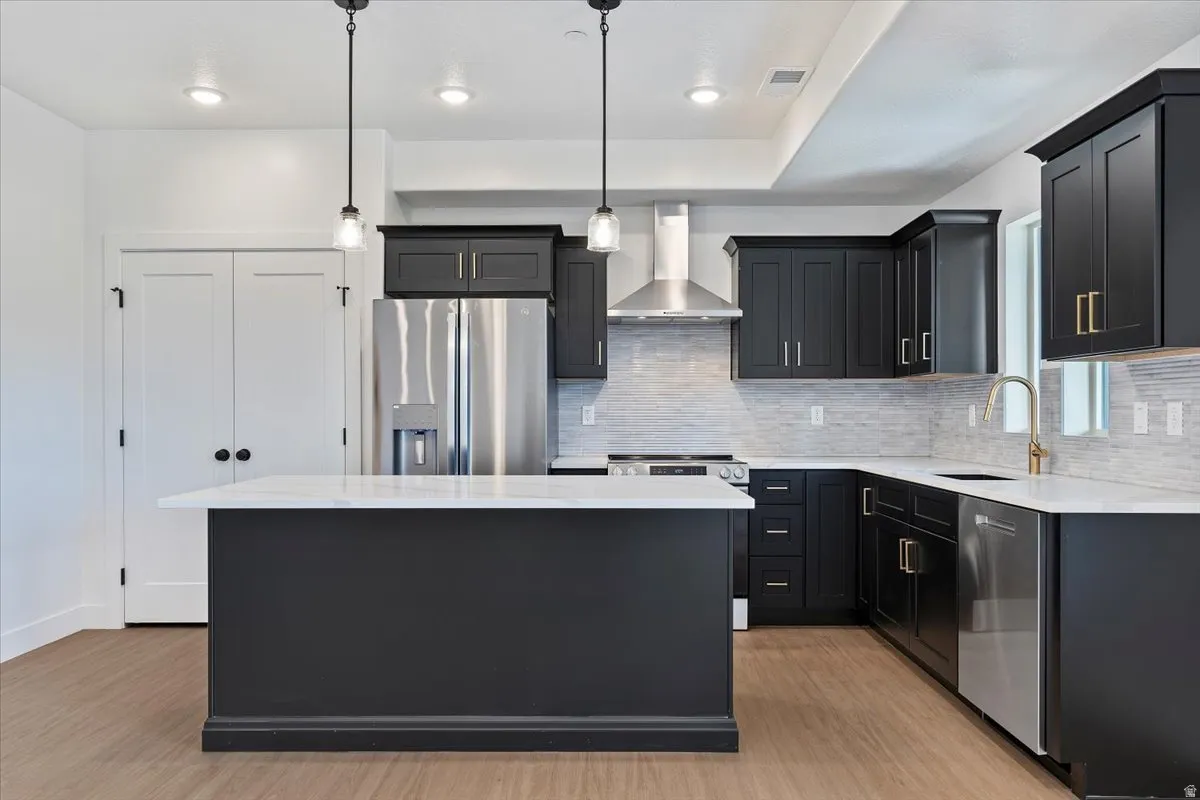 Kitchen featuring dark cabinets, stainless steel appliances, wall chimney range hood, light stone countertops, and decorative light fixtures