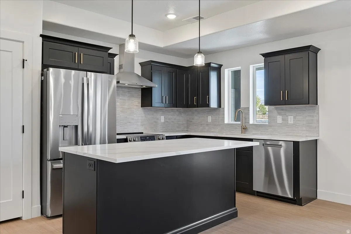Kitchen featuring pendant lighting, dark cabinets, appliances with stainless steel finishes, wall chimney exhaust hood, and backsplash