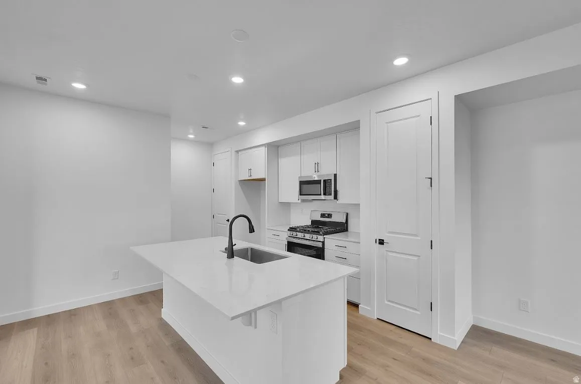 Kitchen featuring appliances with stainless steel finishes, white cabinetry, a kitchen island with sink, light stone countertops, and light wood-type flooring