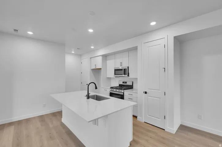 Kitchen featuring appliances with stainless steel finishes, white cabinetry, a kitchen island with sink, light stone countertops, and light wood-type flooring