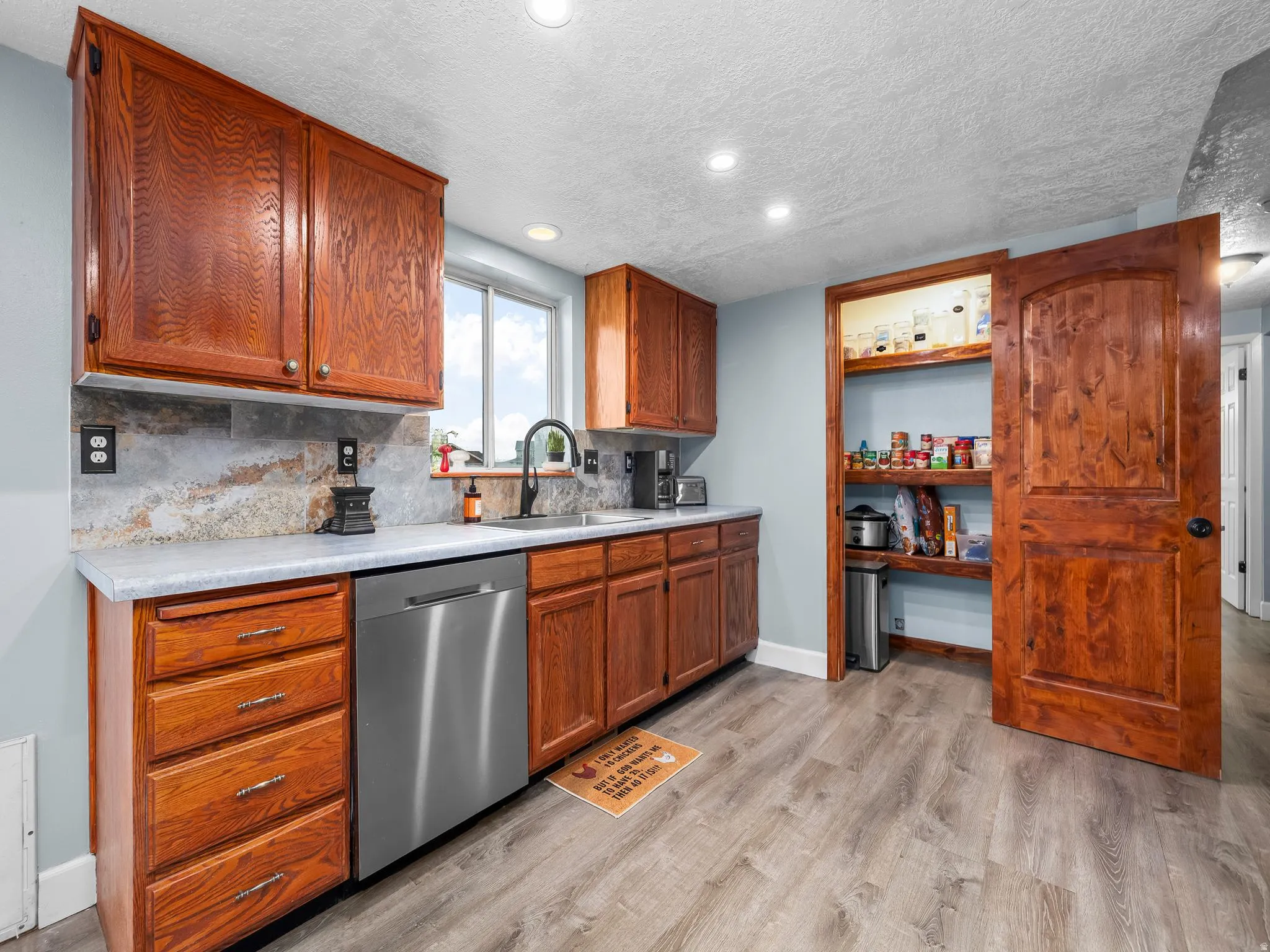 Kitchen featuring light countertops, stainless steel dishwasher, brown cabinetry, a textured ceiling, and decorative backsplash