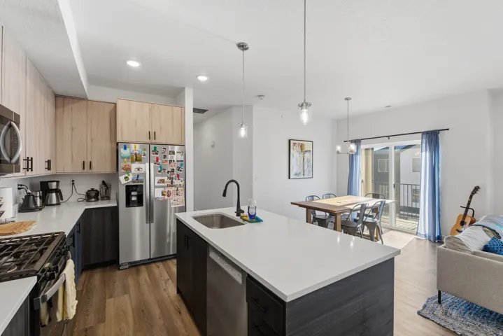Kitchen with stainless steel appliances, hanging light fixtures, an island with sink, dark wood-style flooring, and light brown cabinetry