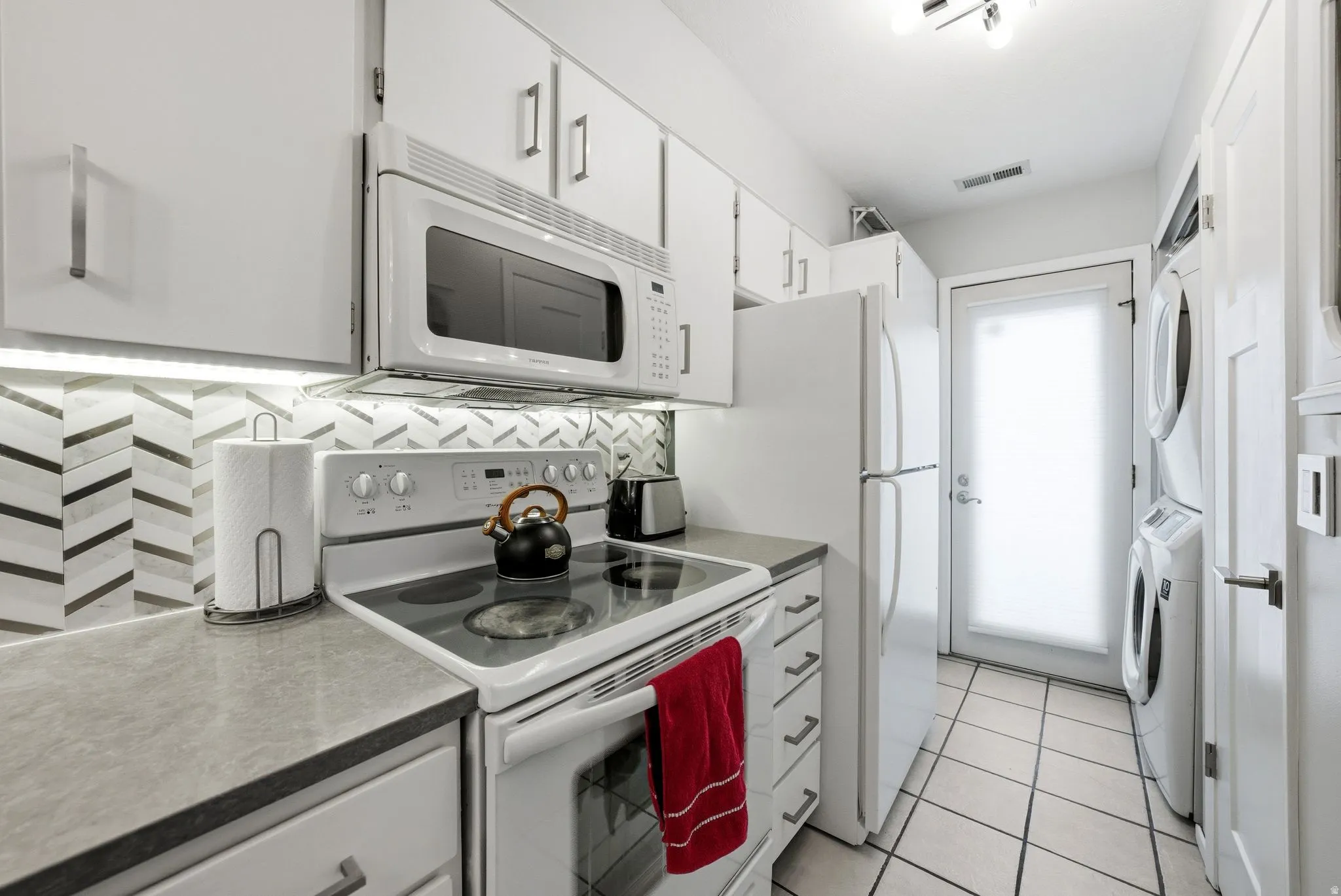 Kitchen with white appliances, white cabinets, light tile patterned flooring, stacked washing machine and dryer, and backsplash