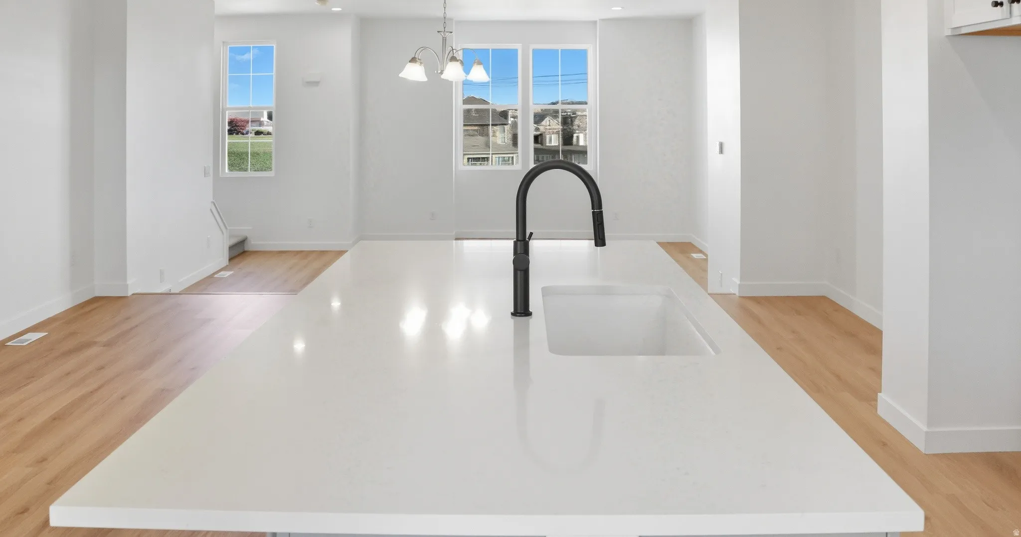 Kitchen view of a center island with sink, hanging light fixtures, light wood-type flooring, light stone countertops, and a chandelier
