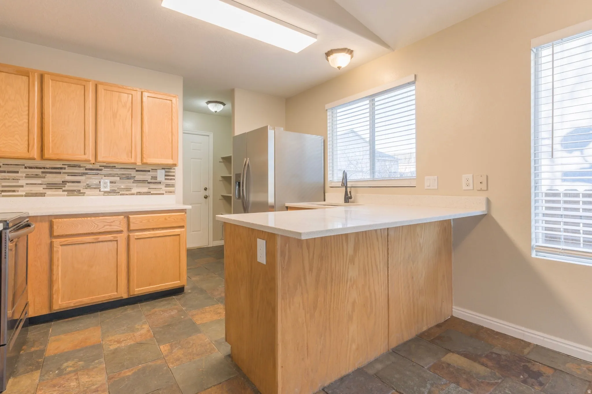 Kitchen with a peninsula, stainless steel appliances, tasteful backsplash, light brown cabinets, and stone tile flooring