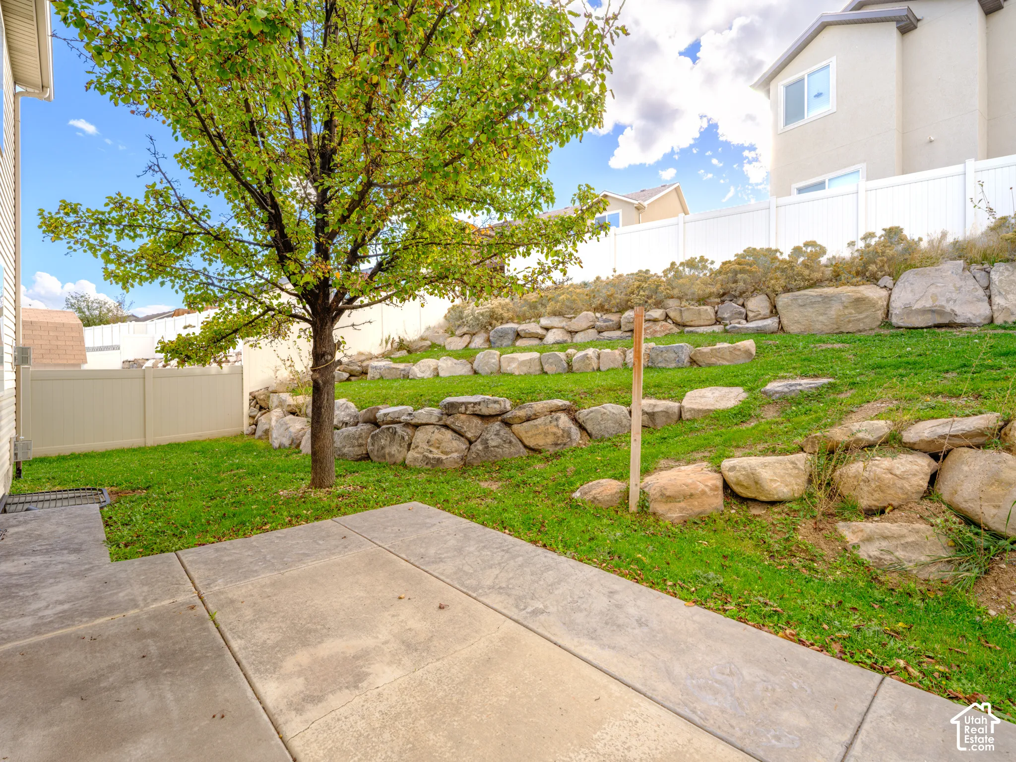 Rear yard featuring a concrete patio, tiered stone retaining walls, lawn area, and white vinyl privacy fencing
