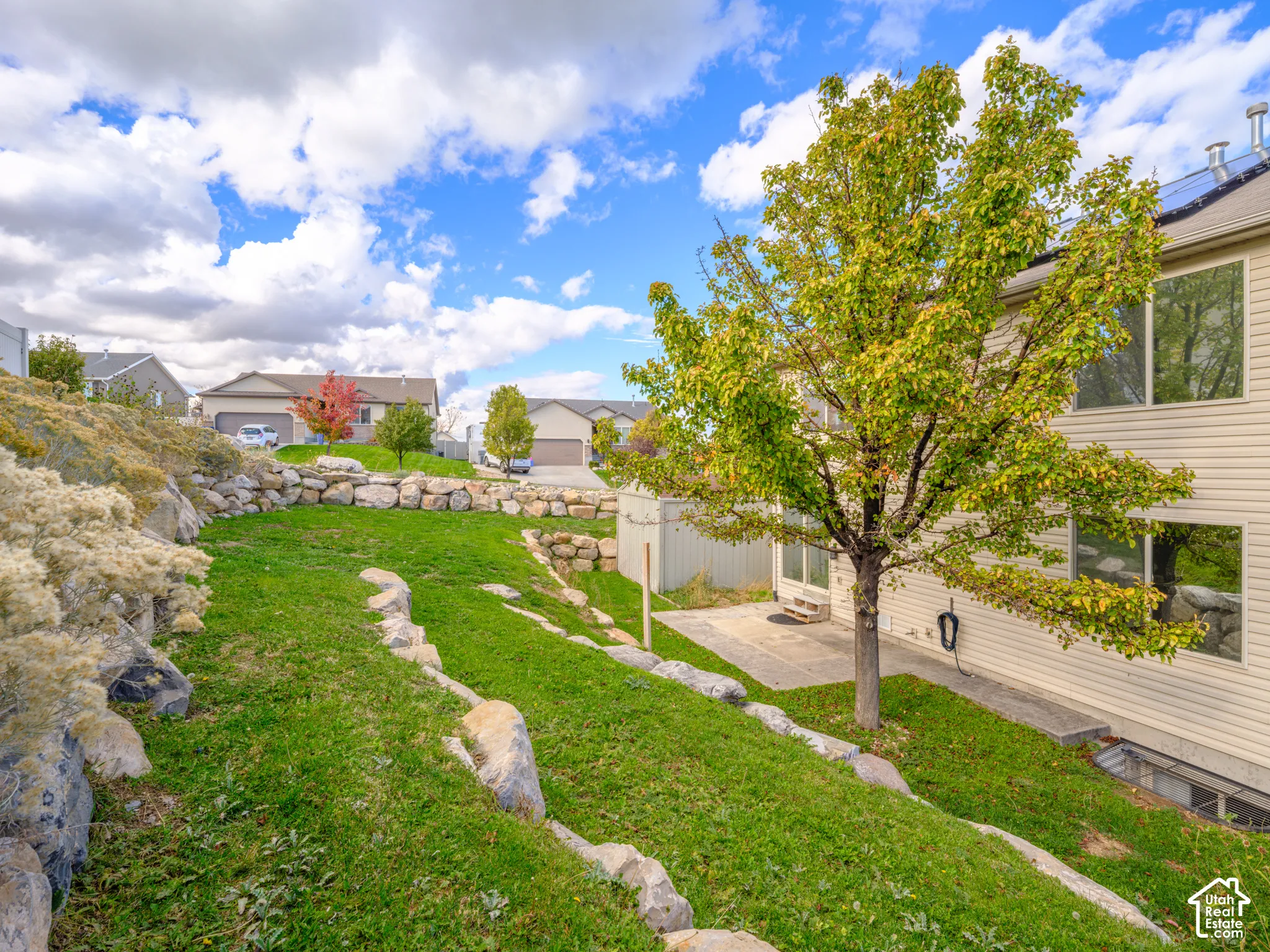 Tiered backyard featuring natural stone retaining walls, manicured lawn, mature tree, and concrete patio