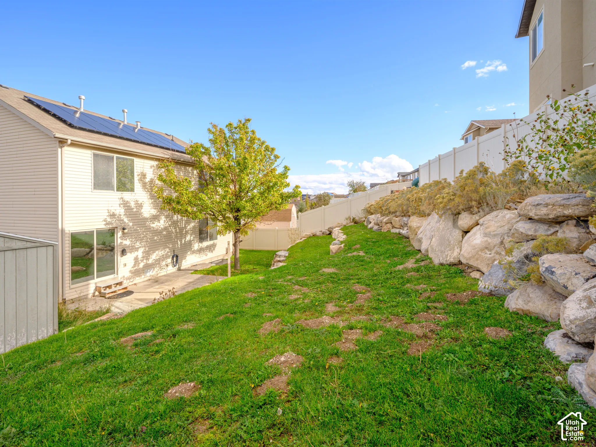 Exterior of the residence featuring roof-mounted solar panels, tiered natural rock landscaping, and a vinyl privacy fence