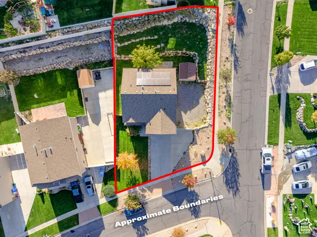 Aerial view of corner lot featuring a concrete driveway, rooftop solar panels, tiered stone retaining walls, and landscaped lawn areas