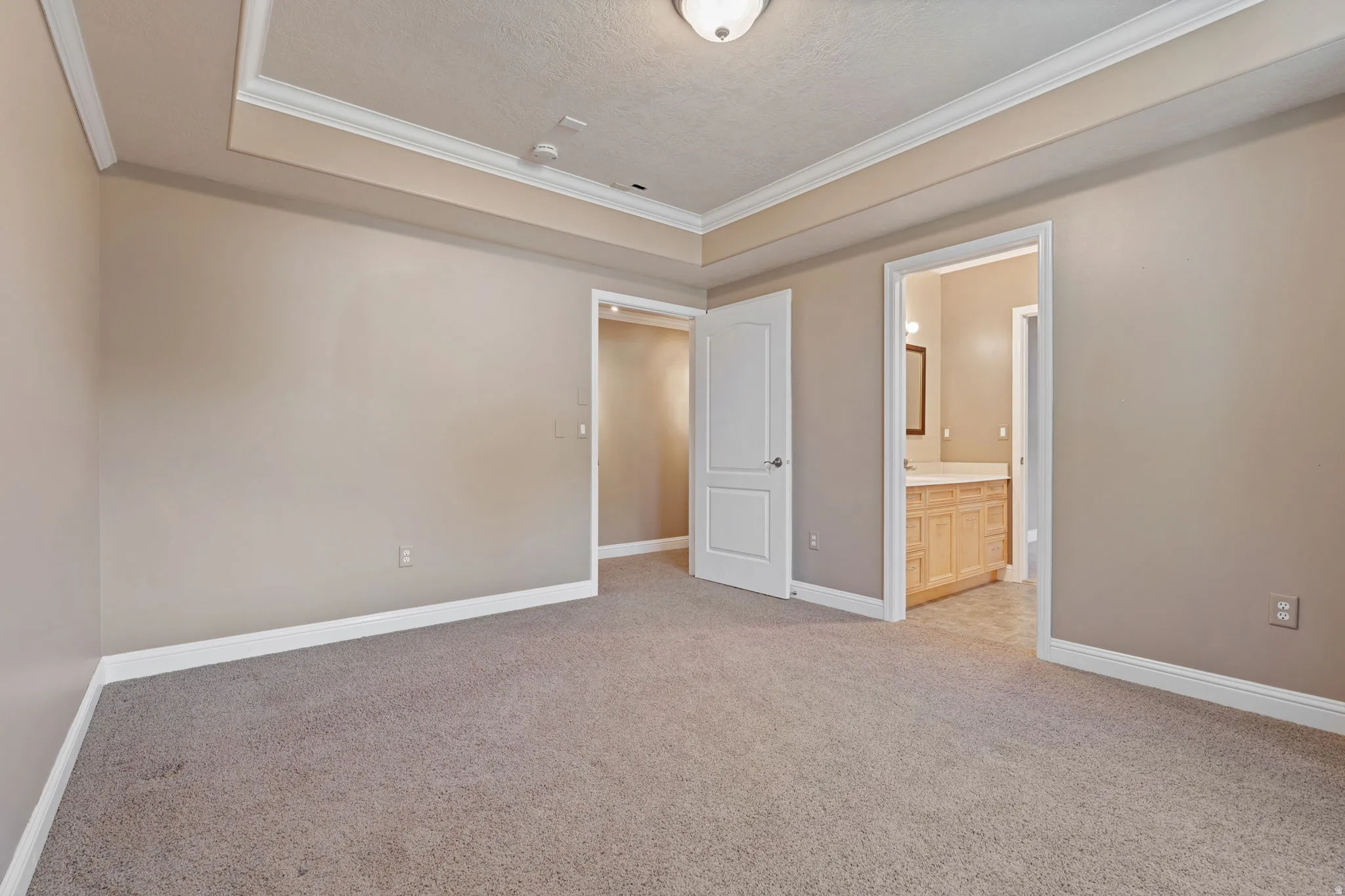 Unfurnished bedroom with light colored carpet, a raised ceiling, crown molding, ensuite bathroom, and a textured ceiling